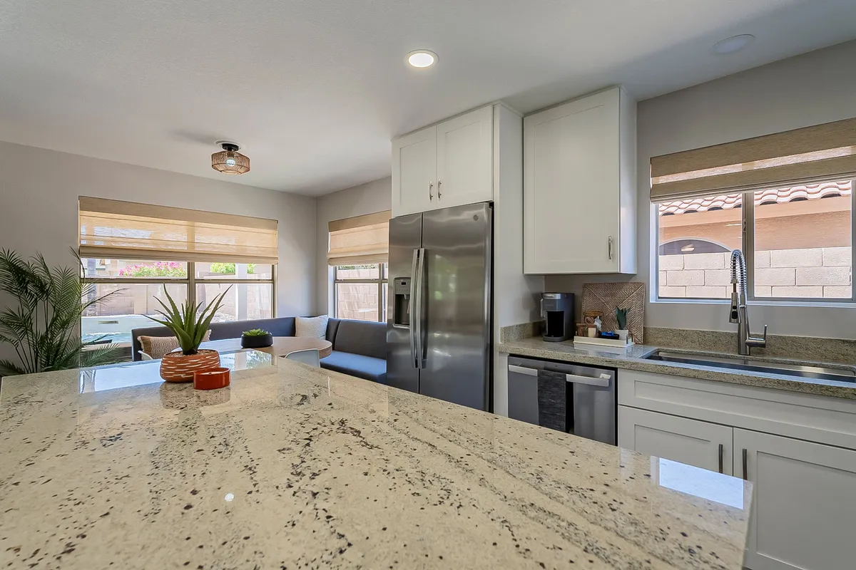 This image features a granite kitchen island as the focal point, complemented by stainless steel appliances and abundant natural light streaming through the windows. A nearby plant decoration and a glimpse of a couch area create a sense of a warm, multi-functional space perfect for cooking, entertaining, or relaxing with family.