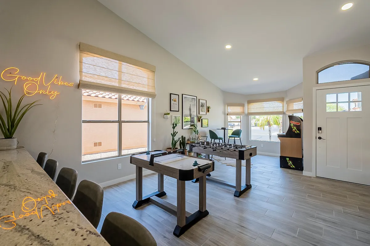 A vibrant, sun-filled game room featuring a foosball table, a neon 'Good Vibes Only' sign, a dining area, and large windows flooding the space with natural light. The scene radiates a cheerful, energetic atmosphere ideal for friendly family tournaments and togetherness.