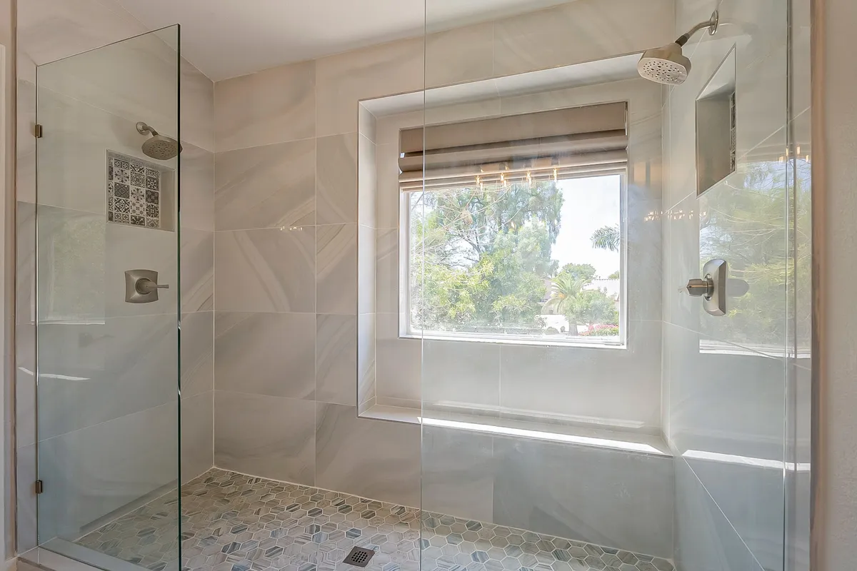 A beautifully lit bathroom featuring a glass shower enclosure, natural light pouring in from a window, and a decorative tile panel. The scene is refreshing and spa-like, perfect for relaxation and self-care.