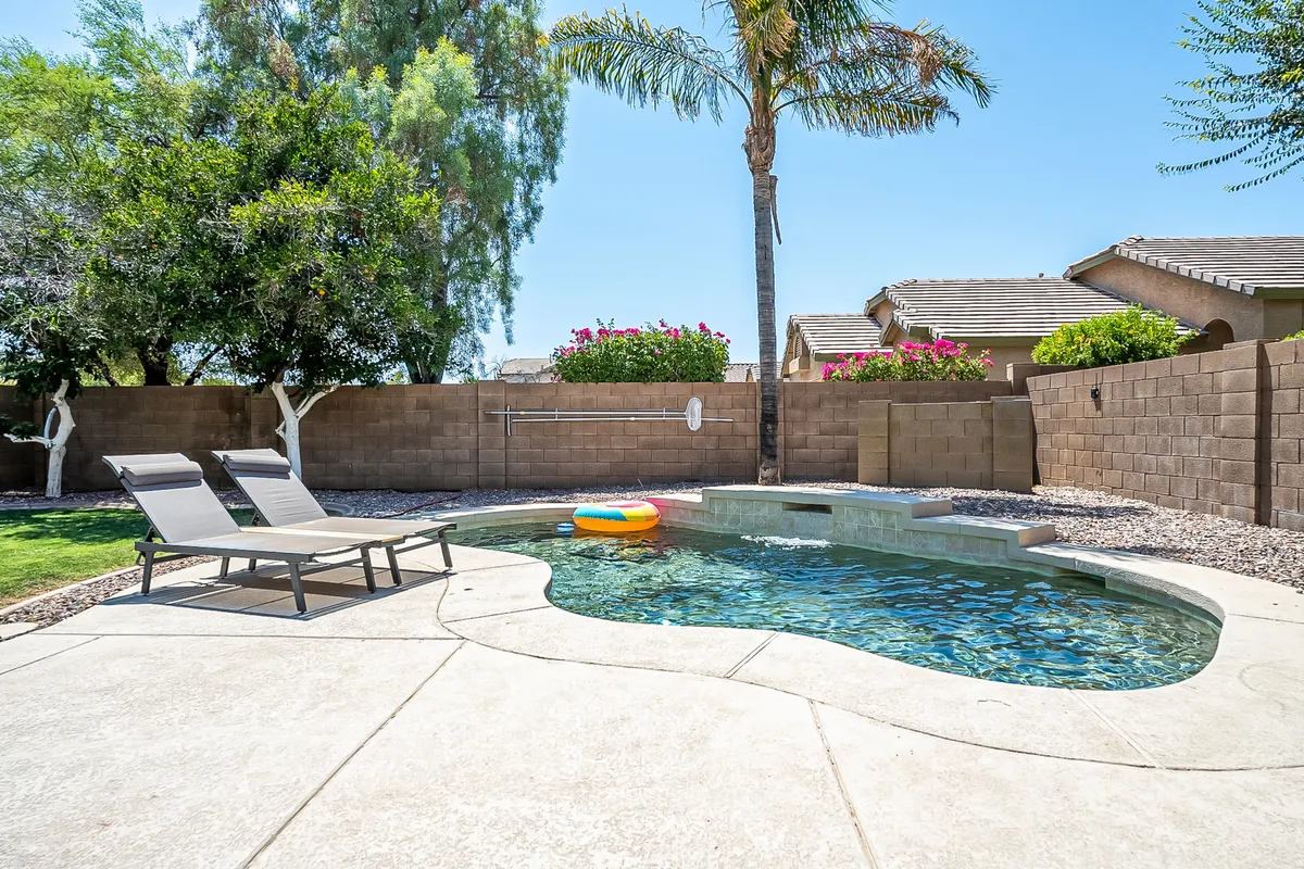 A sparkling swimming pool surrounded by lounge chairs and flowering plants, with a tall palm tree casting shade. The setting is vibrant and lush, ideal for group gatherings and summer fun. The pool area invites sunbathing, swimming, and casual socializing, with plenty of space for friends to gather.