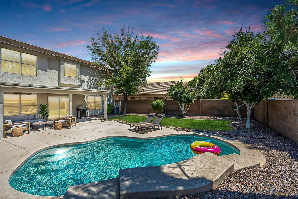 The image showcases a sparkling swimming pool surrounded by lush greenery, with inviting outdoor seating arranged for relaxation and al fresco dining. The warm Gilbert sunset casts a golden glow over the peaceful backyard oasis, hinting at tranquil mornings with coffee and lively summer barbecues by the pool.