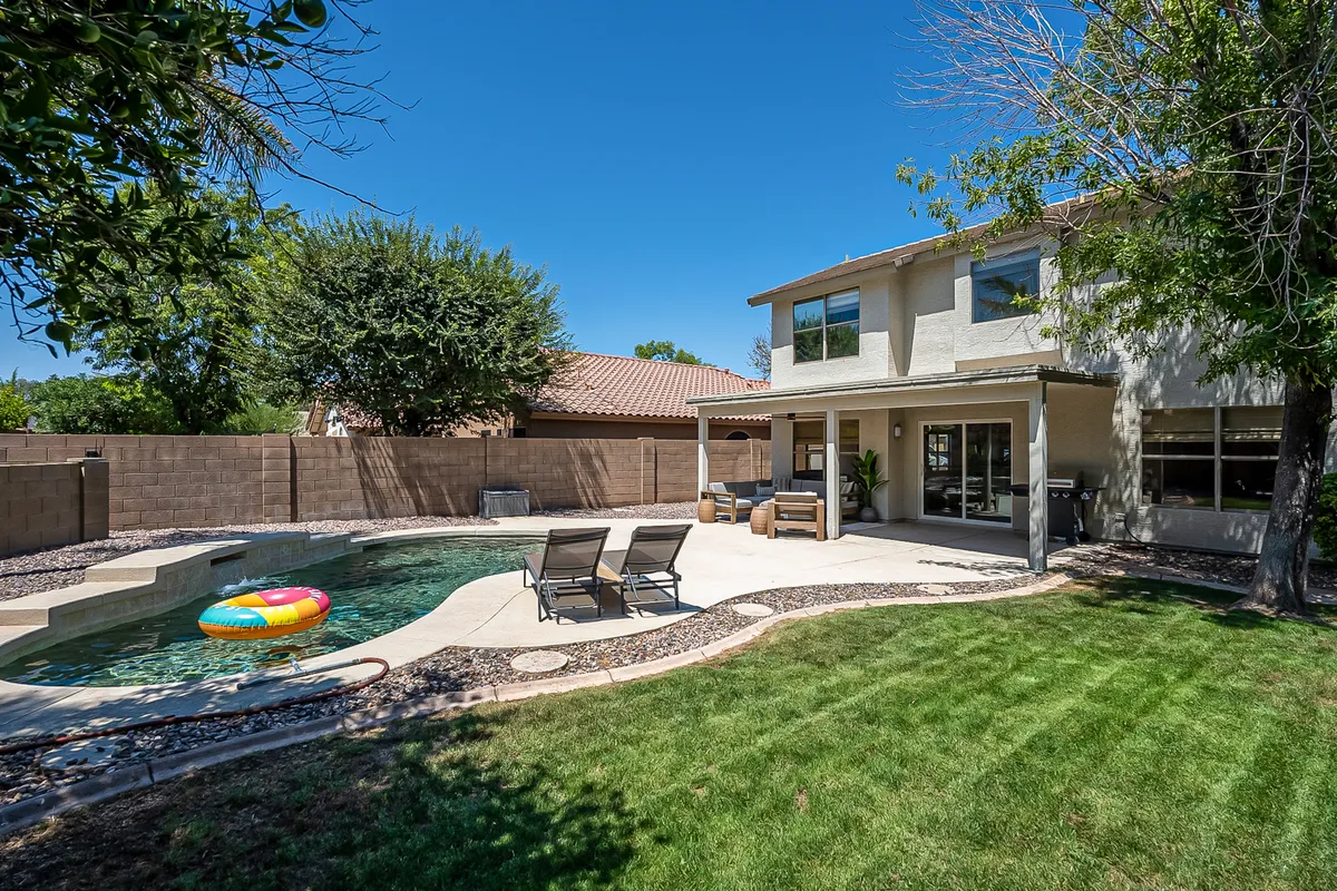 A sparkling swimming pool is surrounded by lounge chairs and a colorful pool float, with a well-maintained lawn and a spacious patio area. The setting is bright, cheerful, and clearly designed for relaxation, play, and outdoor family gatherings.