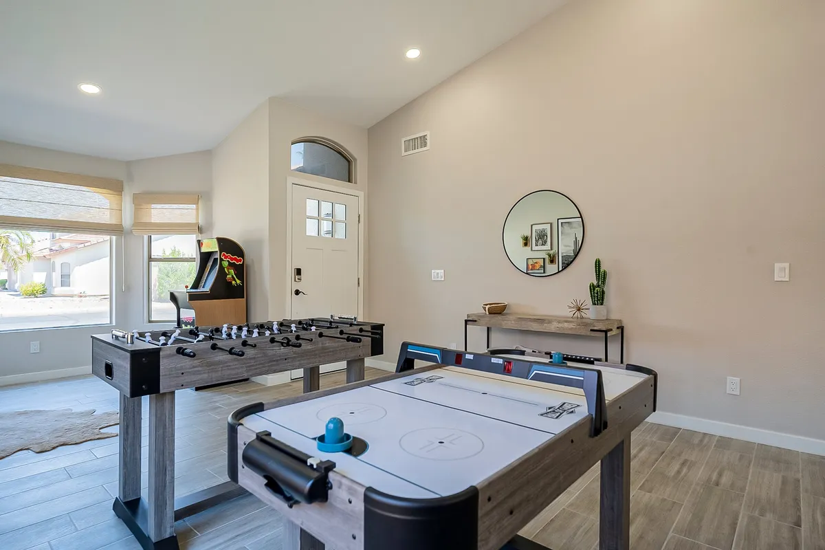 This lively interior image spotlights both a foosball table and an air hockey table set up in a spacious, bright room. An arcade game is also visible along with a circular mirror and a small plant on a side table, capturing the essence of a multifunctional entertainment zone. The layout suggests ample space for active games, group gatherings, and casual relaxation.