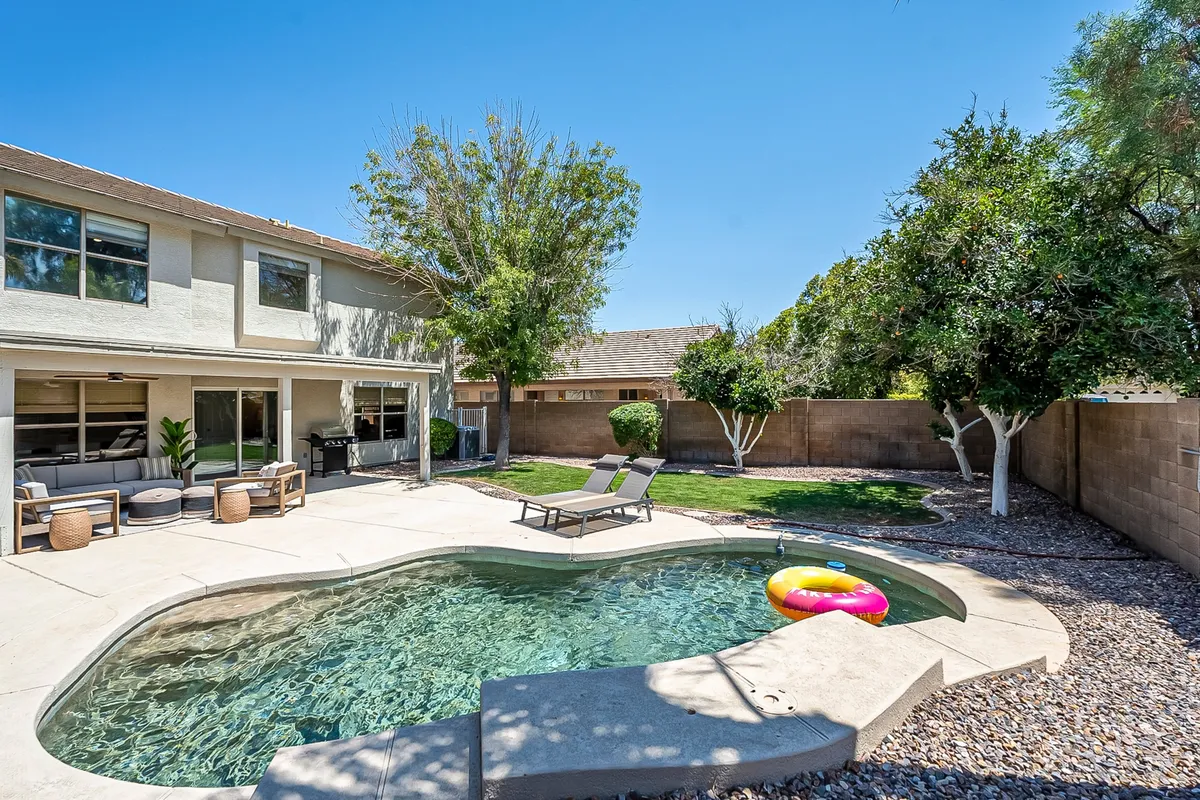A spacious backyard with a shimmering swimming pool, sun loungers, an outdoor seating area, and a lush green lawn bordered by mature trees. The setting radiates relaxation and offers multiple areas for swimming, sunbathing, and dining outdoors.