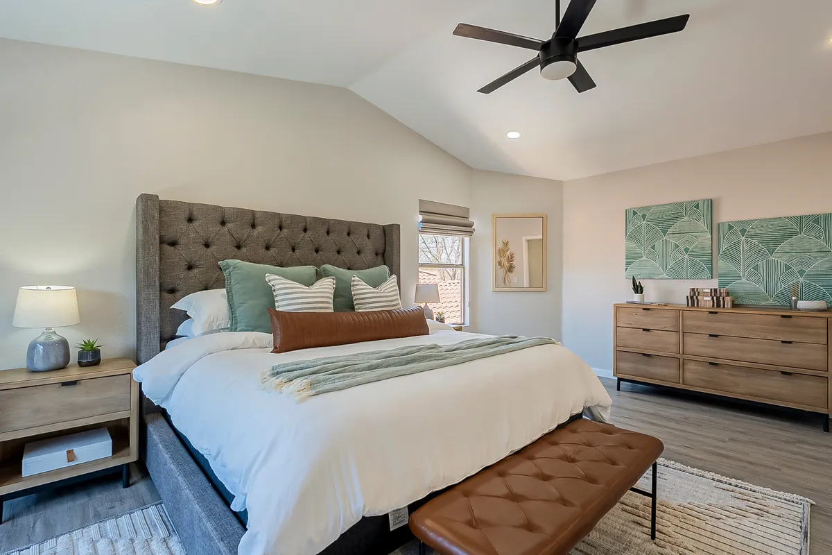 A serene master bedroom featuring a king-sized bed with a tufted headboard, decorative cushions, a wooden dresser, wall artwork, and a bench at the foot of the bed. Sunlight streams in, creating a tranquil and inviting retreat.
