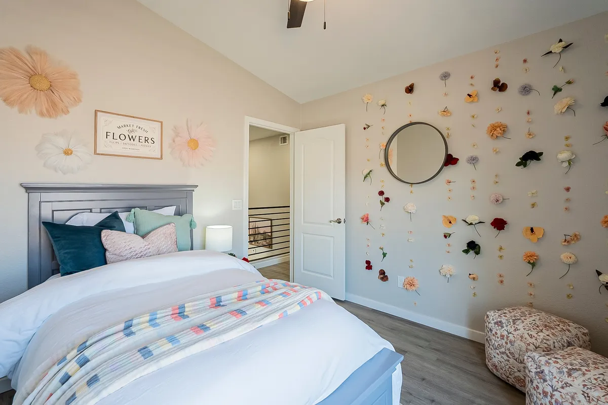 A guest bedroom with a bed adorned in a patterned throw, playful flower wall decor, a round mirror, and an inviting accent chair. The space is airy, youthful, and filled with soft natural light, highlighting a cozy retreat for guests.